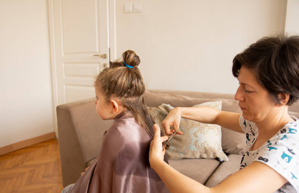 若い女性はパンデミックの最中に娘の髪を自宅で切っています。家にいて. - 写真・画像