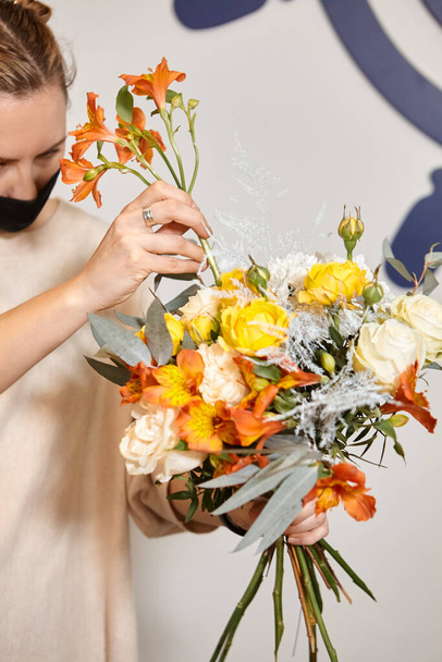 Florist works with colors. Flower seller chooses flowers for future bouquet. Flowers shop worker in a mask standing in flower shop and checking flowers in glass vase. - Photo, Image