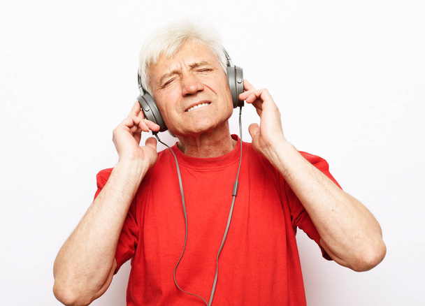 Lifestyle, tehnology and old people concept: elderly gray-haired man witj closed eyes wearing red t-shirt listening to music with headphones over white backgrund - Фото, зображення