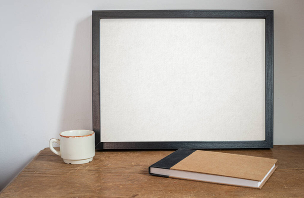cadre noir maquette, sur une vieille table et mur blanc, avec carnet de croquis vierge ou journal et tasse à café .on table en bois . - Photo, image