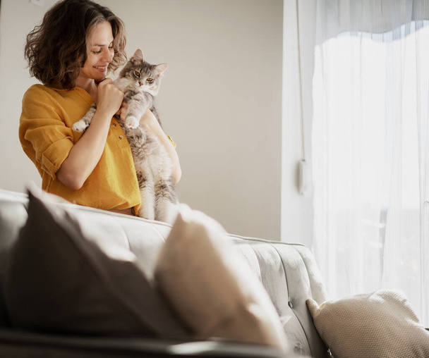 Beautiful cheerful young woman with a cute gray cat in her arms at home in the interior, love for pets - Fotografie, Obrázek