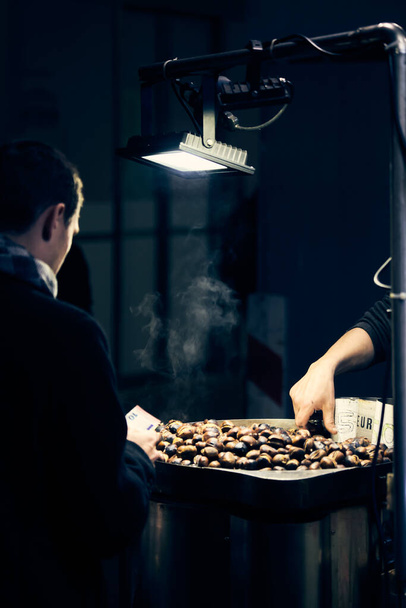 Chestnut seller on the street and man buying grillled chestnuts. Milan, Italy - December 25, 2019 - 写真・画像