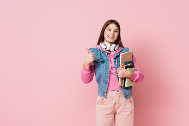 Smiling teenager in headphones showing like and holding notebooks on pink background - Fotoğraf, Görsel