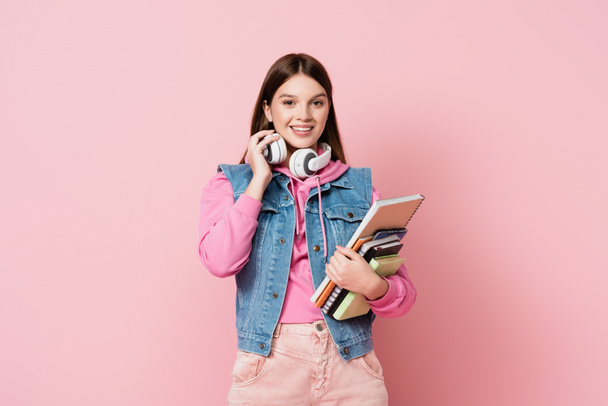 Cheerful girl holding headphones and copy books on pink background - Photo, Image