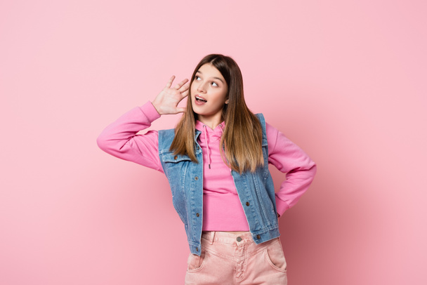 Adolescente con la mano cerca de la oreja mirando hacia otro lado sobre fondo rosa - Foto, imagen