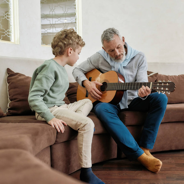 Little boy grandson watching how grandad playing on acoustic guitar while sitting together on sofa in the living room, spending weekend at home - Photo, Image