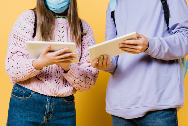 Vue recadrée d'une adolescente dans un masque médical tenant une tablette numérique près d'un ami sur fond jaune - Photo, image