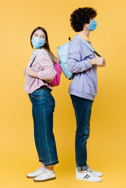 Zijaanzicht van tieners in medische maskers met rugzakken geïsoleerd op geel - Foto, afbeelding