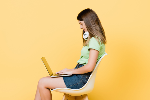 Vista lateral de adolescente en auriculares usando portátil sobre fondo amarillo - Foto, Imagen