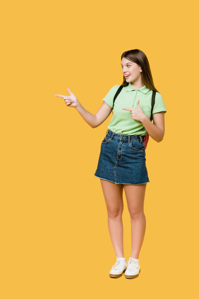 Adolescente sonriente con mochila apuntando con los dedos aislados en amarillo - Foto, Imagen