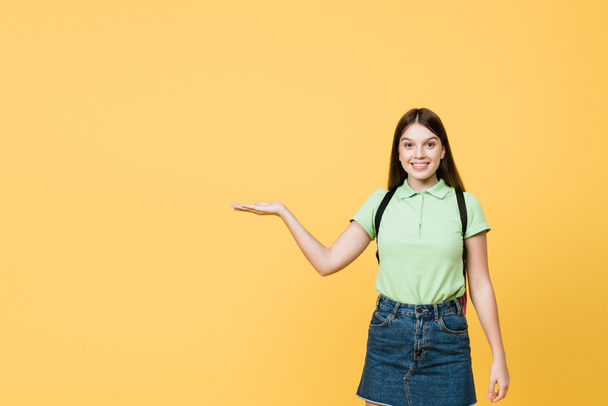 Morena adolescente chica con mochila apuntando con la mano aislada en amarillo - Foto, Imagen