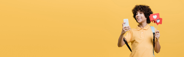Scolaro sorridente che tiene bastoni con cuori di carta e utilizza smartphone isolato su giallo, banner  - Foto, immagini