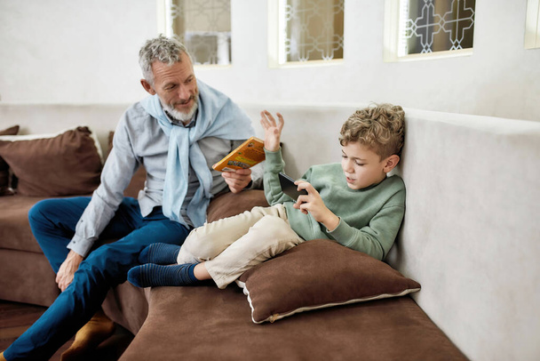 Grandfather giving book to his little grandson while child playing video games on smartphone, they sitting on sofa at home - Φωτογραφία, εικόνα