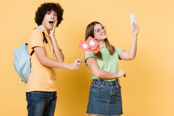 Emocionado adolescente sosteniendo corazones de papel en palos cerca de un amigo sonriente tomando selfie sobre fondo amarillo - Foto, imagen