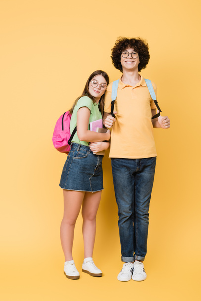 Adolescente sonriente con mochila de pie cerca de un amigo con cuadernos sobre fondo amarillo - Foto, Imagen