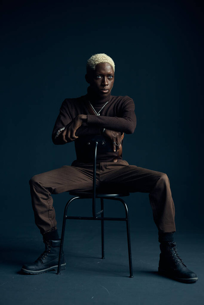 dark-skinned handsome guy with white hair and blue eyes sitting on a chair in a dark studio with a serious expression in brown clothes and silver accessories, he looks into the camera  - Photo, image