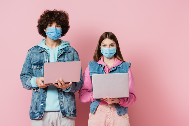 Adolescentes con máscaras médicas mirando a la cámara mientras sostienen computadoras portátiles sobre fondo rosa - Foto, Imagen