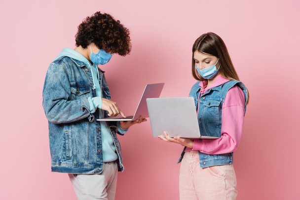 Adolescentes con máscaras protectoras usando computadoras portátiles sobre fondo rosa - Foto, imagen