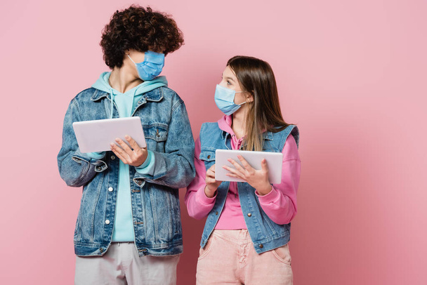 Chica adolescente en máscara protectora sosteniendo tableta digital y mirando a un amigo sobre fondo rosa - Foto, Imagen