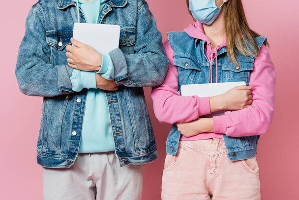 Oříznutý pohled teenagera v lékařské masce drží digitální tablet v blízkosti přítele na růžovém pozadí - Fotografie, Obrázek
