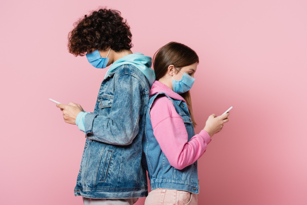 Side view of teenagers in denim jackets and medical masks using smartphones on pink background  - Foto, Imagen