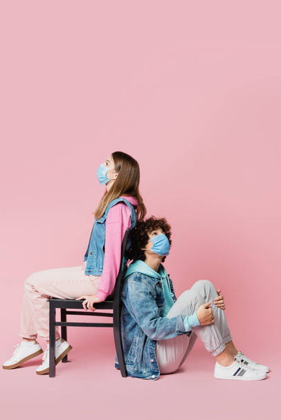 Teenage boy in medical mask sitting near friend on chair on pink background  - Foto, Imagem