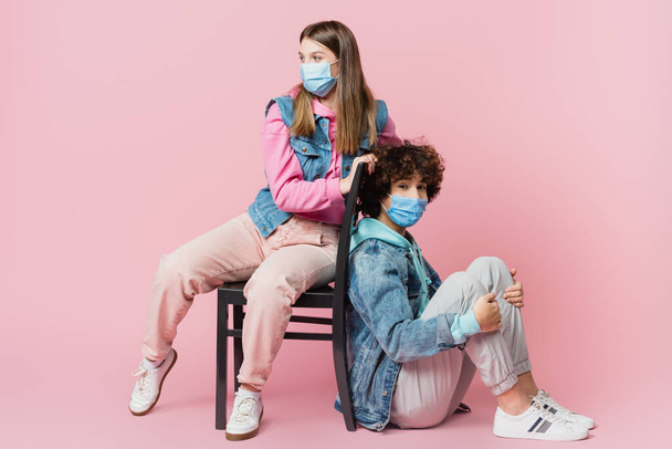 Curly teenager in medical mask looking at camera near friend on chair on pink background  - Photo, Image