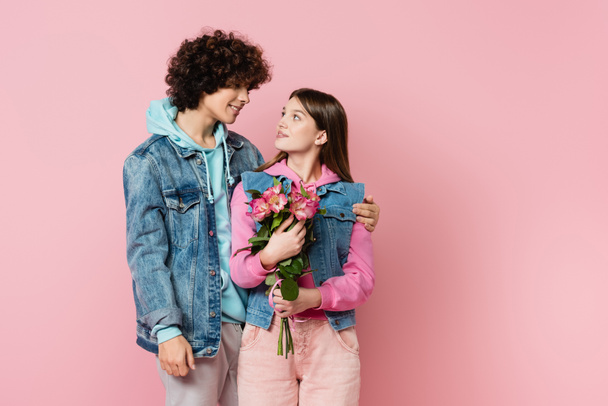 Smiling teenager hugging girlfriend with bouquet isolated on pink  - Фото, изображение