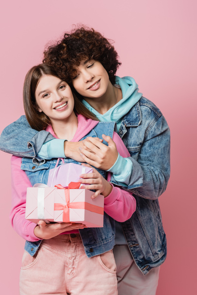 krullend tiener knuffelen glimlachen vriendin met presenteert geïsoleerd op roze  - Foto, afbeelding