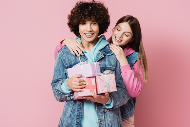Adolescente rizado sosteniendo cajas de regalo cerca de novia sonriente sobre fondo rosa  - Foto, Imagen