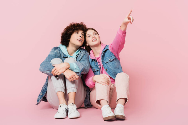 Lächelndes Teenie-Mädchen, das mit dem Finger in der Nähe seines Freundes auf rosa Hintergrund zeigt - Foto, Bild