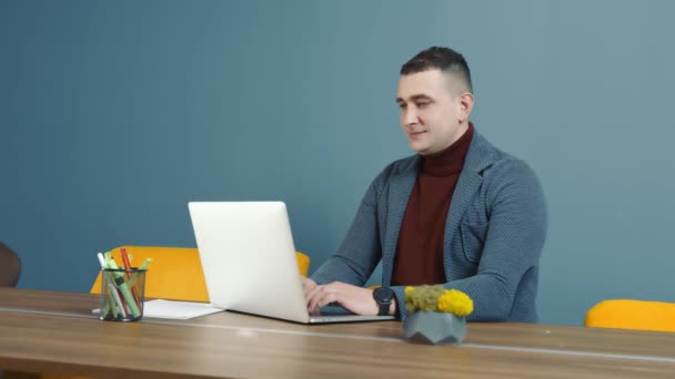 Egy irodai munkás portréja, aki a kamerába néz és mosolyog. Egy férfi ül egy számítógépnél az irodában és dolgozik. Férfi kaukázusi üzletember távolról dolgozik laptoppal. - Felvétel, videó
