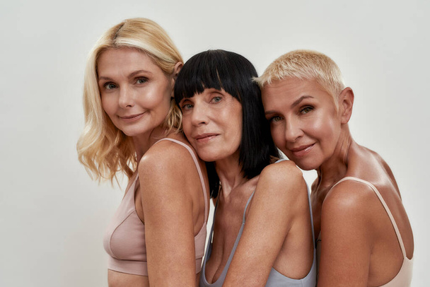 Close up portrait of three beautiful happy mature women in underwear posing together isolated over light background - Photo, Image