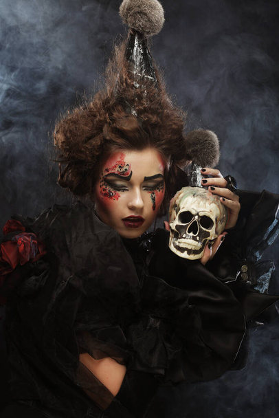 Picture a beautiful fantasy woman with creative make up and hairstyle holding a skull. Halloween theme. Party theme. Over dark background. - Foto, Bild