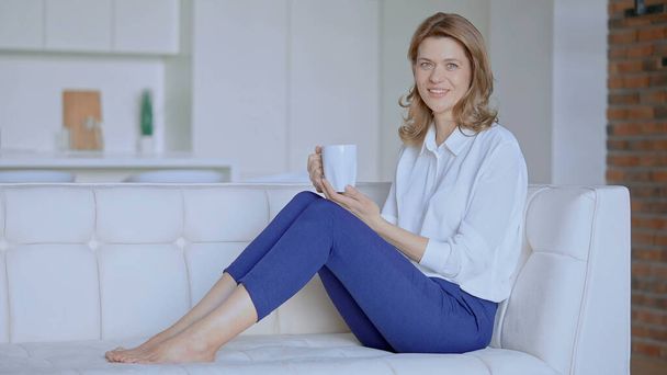 Μια γυναίκα κάθεται στον καναπέ με ένα φλιτζάνι καφέ. - Φωτογραφία, εικόνα