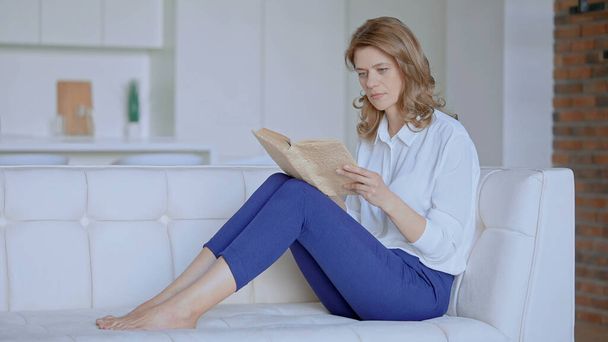 Μια γυναίκα διαβάζει ένα βιβλίο που κάθεται στον καναπέ. - Φωτογραφία, εικόνα