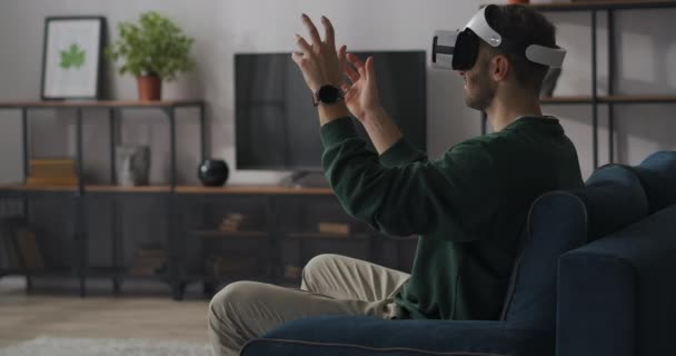 hombre está usando gafas vr en casa, sentado en la habitación y gesticulando por las manos, moviendo objetos virtuales en el aire - Imágenes, Vídeo