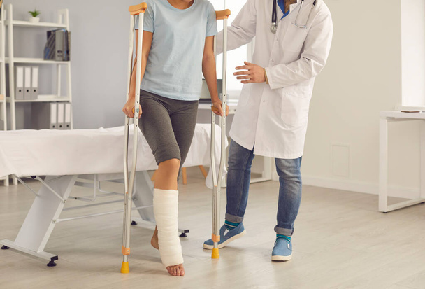 Patient with broken leg walking with crutches assisted by doctor during visit to hospital - Фото, изображение