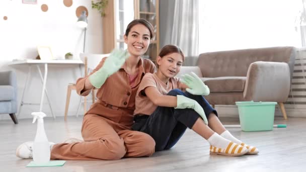 Wide shot of young mixed-race woman and her teenage daughter both wearing rubber gloves sitting on floor in living boom, looking at camera and waving hands before cleaning their house - Video, Çekim