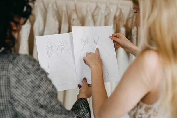 Modedesignerin zeigt einer Kundin in ihrem Atelier Skizzen für Hochzeitskleider. - Foto, Bild