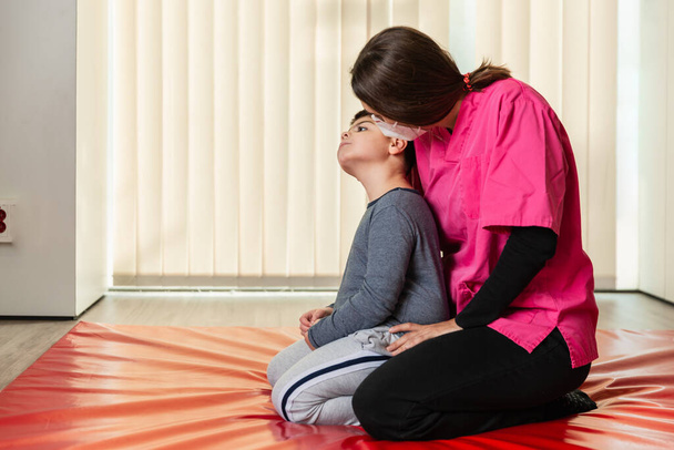 инвалид ребенок и физиотерапевт на красном гимнастическом коврике делает упражнения. пандемическая защита маски - Фото, изображение