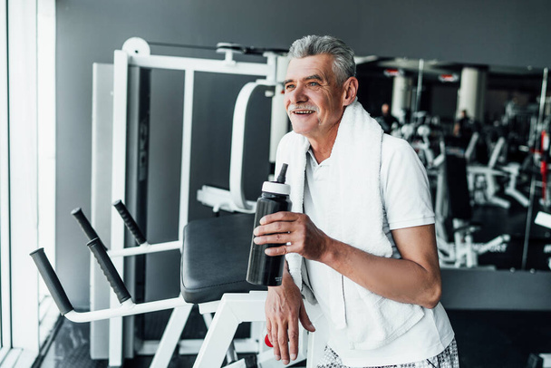 Photo of an older man in the gym during training, he is resting and drinking water. 60+ Healthy appearance, sports lifestyle. - Photo, Image