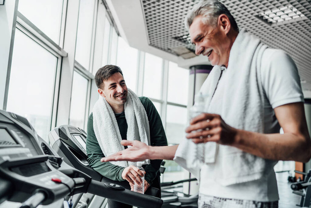 Portrait of two men in the gym, they communicate with each other on treadmills and drink water. Cardio, sports, health. - Fotoğraf, Görsel