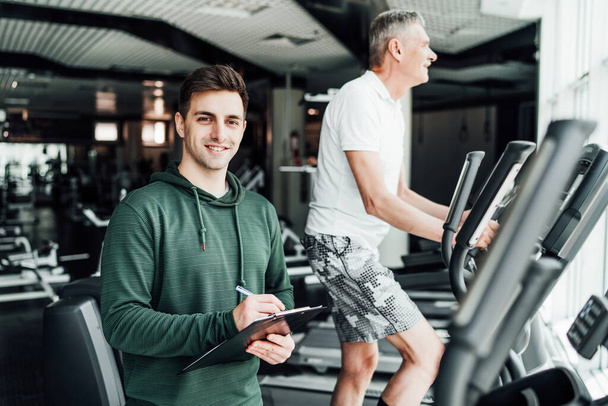 Personal trainer. In the photo, two men, the focus is a good young trainer, he holds a training plan in his hands, on the back of the man performs a cardio exercise. Workout plan, modern gym. - Photo, Image