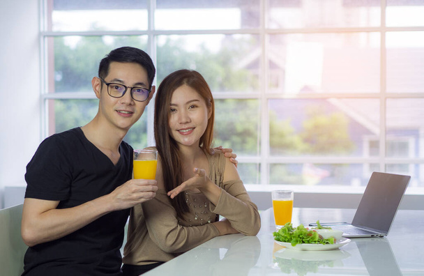 Asyalı genç bir çift, sebze salatası, dizüstü bilgisayar ve mutfakta otururken masaya yerleştirilmiş bir bardak taze portakal suyu tutuyor. Aşk çifti kavramı. - Fotoğraf, Görsel