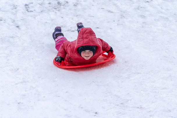 Ein kleines Mädchen fährt auf einem Schlitten von einer Winterrutsche. Winterurlaub. - Foto, Bild