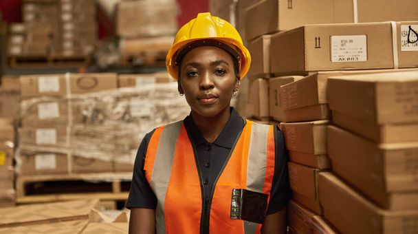 若いアフリカの女性の倉庫労働者は、ボックスのスタックに対して傾いて安全ベストとヘルメットを着て - 写真・画像