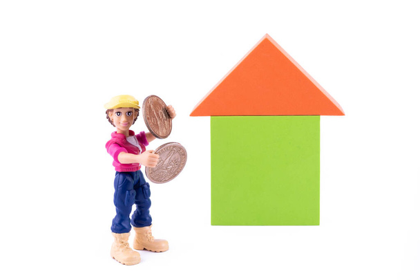 Une figurine en plastique symbolique d'un homme tenant des pièces de monnaie en coupures de 1 dollar américain près d'une maison conventionnelle faite de cubes d'enfants sur fond blanc - Photo, image