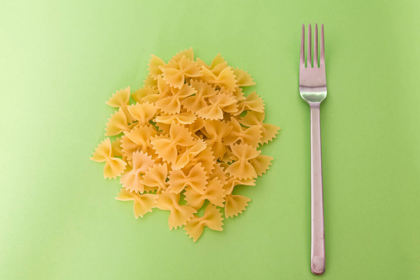 Pile of raw dough in the shape of a bow tie is resting on a green background with a steel fork nearby. - Foto, Imagem