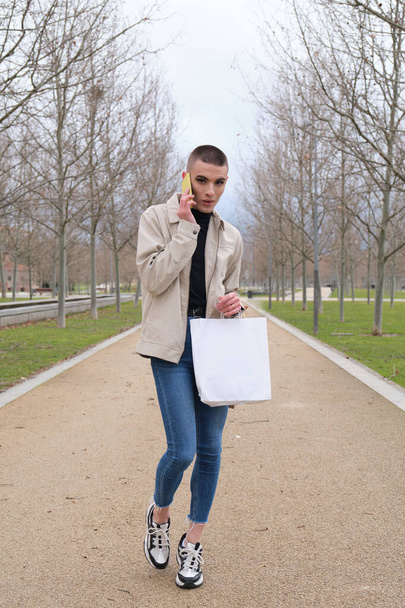 Όμορφος νεαρός άνδρας που φοράει μακιγιάζ, περπατά και μιλάει στο smartphone του, κουβαλώντας τσάντες για ψώνια. Μη δυαδικός ανδρόγυνος τύπος. - Φωτογραφία, εικόνα